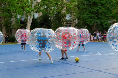 Mavi sahada, şişme topların içindeki oyuncuların sarı bir topu kovaladığı enerjik balon futbol maçı. Seyircilerin izlediği eğlenceli bir açık hava aktivitesi.