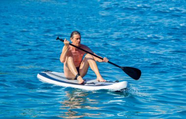 50 'li yaşlarda orta yaşlı bir adam denizde ayakta duran bir kürek tahtasında denge kurmaya çalışıyor, bazen oturuyor ve bazen de suya düşüyor. SUP tahtasının eğlenceli ve otantik sahnesi.