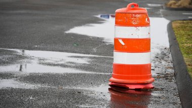 Canlı turuncu ve beyaz bir trafik konisi yolun karanlık, ıslak asfaltına karşı duruyor. Yansıtıcı yüzeyi son yağmurla benekli..
