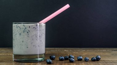 Bir bardak muzlu ve yaban mersinli, pembe pipetli meyve suyu ahşap bir masada etrafına dağılmış yaban mersinleri ile oturur.