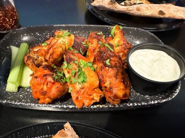 İçinde taze otlarla süslenmiş baharatlı tavuk kanatları ve yanında kereviz çubukları ve mavi peynir sosu bulunan bir tabak.