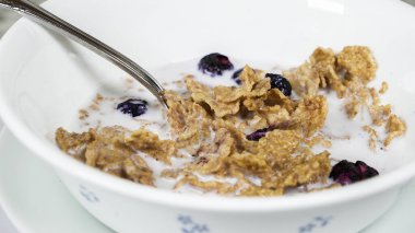 A bowl filled with crunchy cereal and blueberries swimming in milk sits on a dining table. A silver spoon rests inside, ready for a delicious breakfast experience. clipart