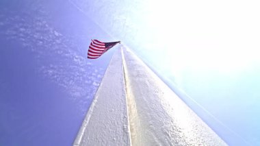 Ön planda uzun bir bayrak direği duruyor, dalgalı Amerikan bayrağı rüzgarda hafifçe dalgalanıyor. Açık mavi gökyüzü vatansever görüntüyü arttırıyor..