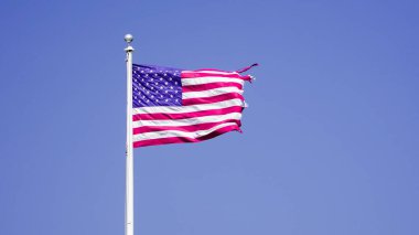 Yukarıda gururla dalgalanan Amerikan bayrağı kırmızı, beyaz ve mavi renkler sergiliyor. Açık mavi gökyüzü, görkemli bir görüntü yaratmak için bir zemin görevi görüyor. Bayrak hafifçe rüzgarda dalgalanıyor..