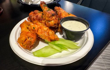 Bir tabak lezzetli, baharatlı tavuk kanatları yanında gevrek kereviz çubukları ve küçük bir kase mavi peynir sosu içerir.