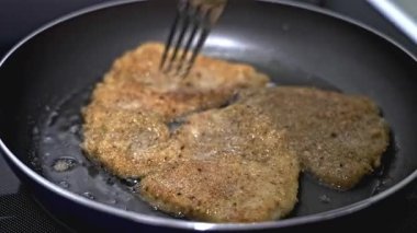 In a kitchen, chicken fillets are being fried in a pan, turning golden brown. A fork is used to flip the fillets, ensuring even cooking and perfect crispiness
