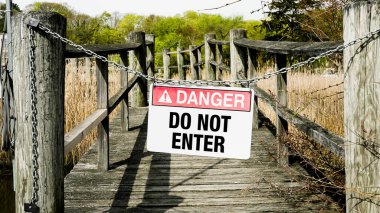 Tahta bir rıhtımın karşısındaki zincirde bir uyarı işareti asılı. Rıhtım arka planda uzun otlar ve ağaçlarla kaplı bataklığa açılıyor.