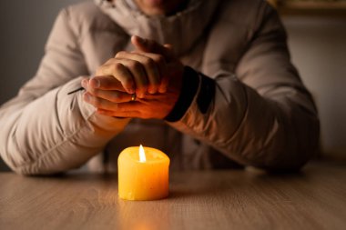 Isıtma ve elektriği kapatın. Sıcak kış ceketi giymiş bir adam evinde bir masada oturur ve yanan bir mumla ellerini ısıtır. Elektrik kesintisi, elektrik kesintisi, yük boşaltımı veya enerji krizi.