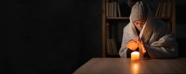 Unhappy woman warm hands on candle at cold home, shutdown of heating and electricity, power outage, blackout, load shedding or energy crisis, concept image. Banner clipart