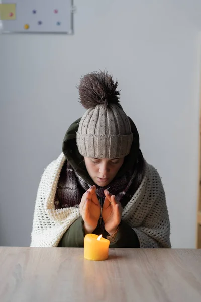Tired sick woman freezing at home, sitting at the table warming hands of burning candle. Woman with home heating problem feeling cold. Concept of no heating in winter at home.