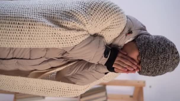 毛布に身を包んだおじさんが寒い寝室に腰を下ろしている 健康を害している若者は家で凍死する 暖房の概念なし 垂直ビデオ — ストック動画