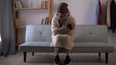Şapkalı ve battaniyeli sağlıksız bir adam soğuk oturma odasında sıcaktan muzdarip. Sağlıksız genç adam evdeki dondurucu soğuktan mücadele ediyor. Isıtma kavramı yok.