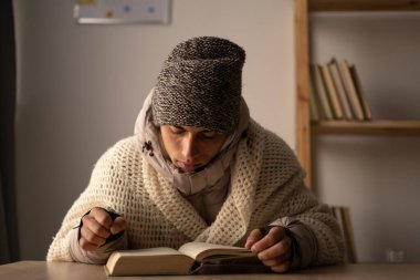 Kışlık giysiler içinde soğuk kitap okuyan bir erkek öğrenci ısıtması olmadan pencerenin kenarında oturuyor. Boşluğu kopyala