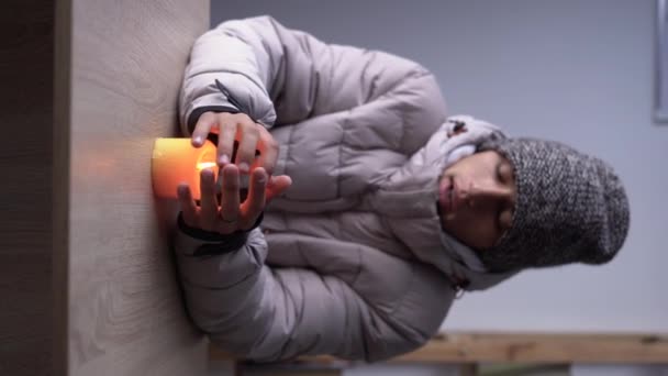 身穿保暖冬帽和夹克的年轻冻人坐在桌旁 用燃烧的蜡烛暖着双手 冬天在家里不能取暖的概念 垂直录像 — 图库视频影像