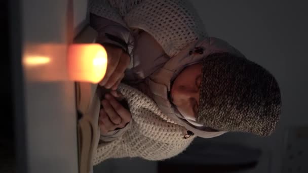 停電と暖房の概念 ヨーロッパのエネルギー危機 冬に家の中で寒さを感じるろうそくの明かりで本を読んでいる若い男 垂直ビデオ — ストック動画