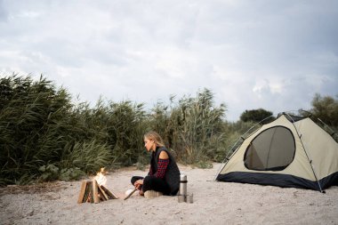 Deniz kıyısında akşam kampı. Genç bayan turist, güzel mavi gökyüzünün altında kamp ateşinin yanında sahilde oturuyor. Turizm ve doğa kavramının güzelliği.