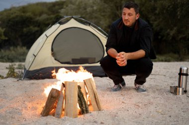 Erkek turist, akşam deniz kıyısındaki kampında dinleniyor. Kamp ateşi ve çadırın yanında oturan adam. Seyahat ve insan konsepti