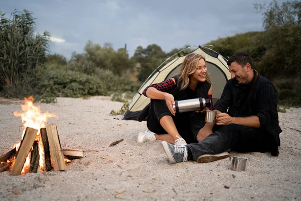 Happy smiling couple traveler sits near a tent by a campfire and pours coffee or tea from a thermos in a seashore in the weekend. Camping, recreation, hiking concept