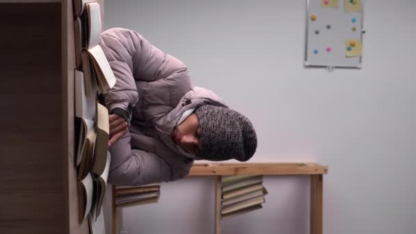 暖かい冬の帽子とジャケットを身に着けている学生は テーブルに家に座って本を勉強します 家庭では冬の暖房の概念 垂直ビデオ — ストック動画