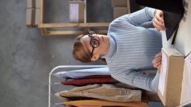 Genç iş kadını ev ofisinin arka planında ürün kutusuyla baş ağrısı ve stres hissediyor. Sağlık ve ofis sendromu kavramı. Dikey video