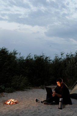 Çadırın yanında dizüstü bilgisayarla oturan yakışıklı bir adam ve alacakaranlıkta kamp yaparken uzaktan kumandayla çalışan bir erkek serbest yazar göl yolculuğunda kamp alanında uzaktan çalışmanın keyfini çıkarıyor.