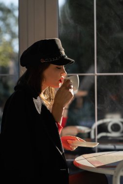 Süslü siyah şapkalı çekici bir kadının portresi ve sonbahar parlak kırmızı süveteri kafenin içinde oturuyor ve harika bir tatil zamanının tadını çıkarıyor. yan görünüm