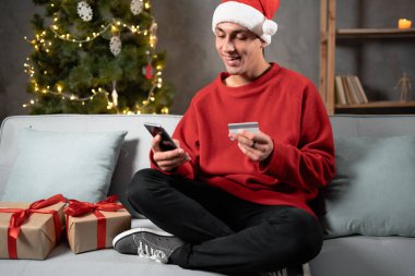 Büyük Noel indirimi, online alışveriş ve satın alma. Noel Baba şapkalı neşeli erkek öğrenci kredi kartı tutuyor ve evdeki kanepede akıllı telefonla ödüyor. Boş boşluk