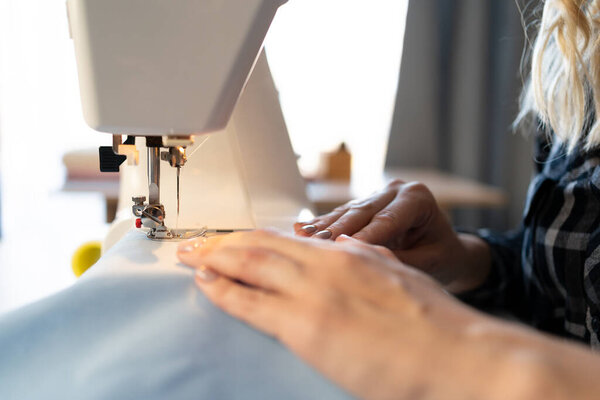 Close up view of sewing process. Female hands stitching blue fabric on professional sewing machine at workplace. blurred background. Copy space