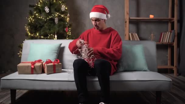 サンタの帽子の怒って悲しい動揺した男は 自宅のソファに座っている間 クリスマスライトが緊張しているのを解明しようとしています クリスマスと人々のコンセプトは — ストック動画