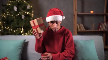 Tahmin et içinde ne var. Noel Baba şapkalı genç adam Noel hediyesini sallıyor, hediye kutusundaki çıngırakları dinliyor koltukta oturuyor. Noel hediyesi konsepti