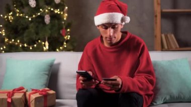 Arap adam Noel hediyelerini kredi kartıyla ödüyor. Akıllı telefon kullanıyor, internetten alışveriş yapıyor, kış tatilinin tadını çıkarıyor. Kanepede şenlik ağacının yanında oturuyor. Noel çevrimiçi indirimi