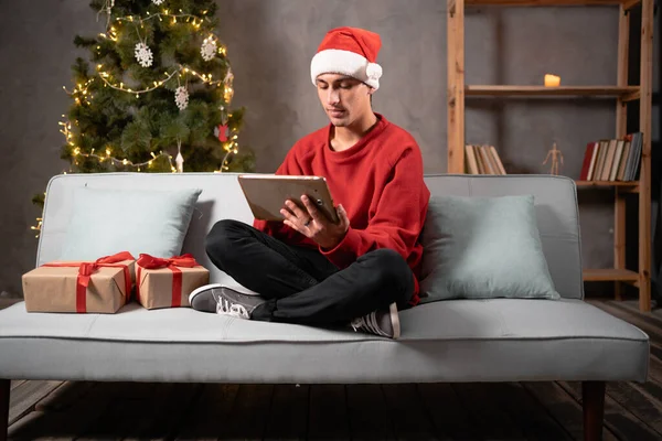 Smiling man sitting on the sofa by the Christmas tree and using tablet. Freelancer working on Holiday xmas. Copy space