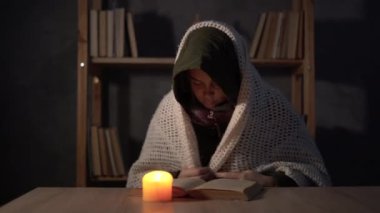 Sıcak kış ceketi giymiş bir kadın evinde bir masaya oturur ve yanan bir mumla ellerini ısıtır. Kışın evde ısıtma ve elektrik olmaması kavramı.