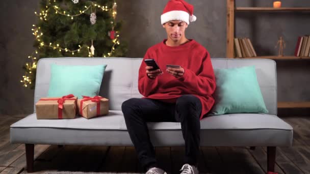 スマートフォンを使用してクリスマスプレゼントのためのクレジットカードでオンラインで支払うサンタハットのアラビア人男性 オンラインショッピング 冬の休日を楽しんで ソファの上にお祝いの木の近くに座って クリスマスセール — ストック動画