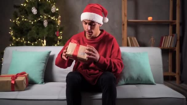 若いアラビア人の男はソファの上の自宅でクリスマスツリーに座って贈り物をアンパック 休日のコンセプト — ストック動画