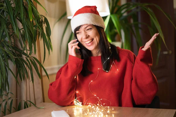 Young smiling happy woman expressing surprise on face and gesturing while speaking on mobile phone. Hipster in Santa at home interior. Christmas call for family. Copy space