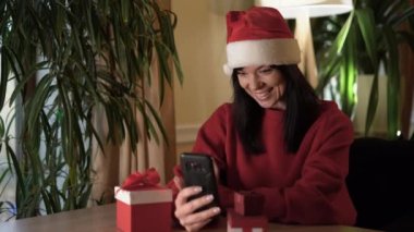 Noel Baba gibi giyinmiş beyaz bir kadın evdeki masada oturuyor ve elinde bir akıllı telefon tutuyor. Video görüşmesi yapıyor. İnternet teknolojileri.
