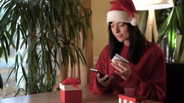 年轻的高加索女人穿着圣塔克洛斯的衣服坐在家里的桌子旁 手里拿着智能手机和信用卡购物 圣诞销售概念 — 图库视频影像