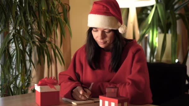 年轻的高加索女人穿着圣塔克洛斯的衣服坐在家里的桌子旁 用笔记本里的笔写字 为圣诞节准备礼物清单的概念 复制空间 — 图库视频影像