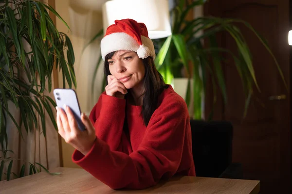 Upset confused woman in Santa hat holding smartphone, looking at mobile phone screen with worried expression, sad female, unpleasant message on Christmas. Bad news concept.