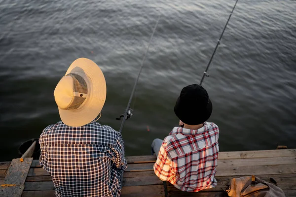 Back view of father and son sitting together on pier fishing with rods in calm lake waters, wearing checkered shirts and hats. Father\'s Day. Copy space