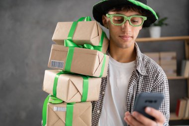 St. patrick's day sale, online shopping man in green leprechaun hat with stack of gift boxes using smartphone. Portrait