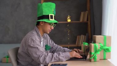 Aziz Patrick Günü 'nde yeşil cüce şapkalı mutlu bir adam 17 Mart' ta elinde hediye kutusuyla evde oturan dizüstü bilgisayarı kullanıyor. İnternet sitesi internet alışverişi ve tatil indirimi. Boşluğu kopyala