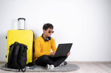 Young guy traveler with a suitcase wears in an orange sweater sits typing on a laptop on isolated white background. freelancer working at the airport. Copy space