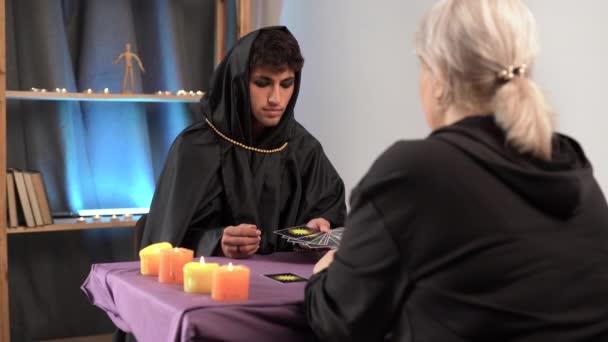 男巫穿着深色哥特式衣服坐在桌旁 艺术服装 算命先生为客户阅读塔罗牌上的未来 祭祀用的蜡烛 场景占卜 — 图库视频影像