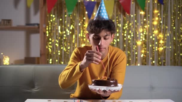 Young Man Cone Hat Putting Candles Cake Celebrating Birthday Home — Vídeo de stock