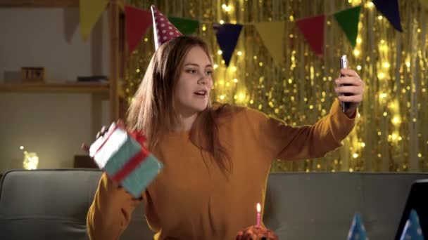 誕生日プレゼントの配達 自宅でビデオ通話でオンラインパーティーを持つ電話の前で誕生日プレゼントを保持する楽しい若い女性 現代の祝日 — ストック動画