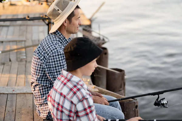 Side view of father and son sitting together on pier fishing with rods in calm lake waters with landscape of setting sun, both wearing checkered shirts. Copy space