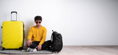 Sarı bavullu tatminsiz yolcu, beyaz arka planda başarısız bir yolculuk yüzünden üzgün oturuyor. Hava yolculuğu konsepti. Pankart