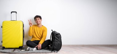 Elinde bavulu olan genç bir Arap, beyaz arka planda iki numarayı parmaklarıyla ya da zafer işaretiyle gösteriyor. Pankart